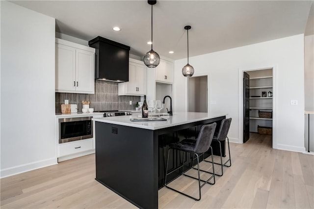 kitchen featuring pendant lighting, built in microwave, white cabinetry, a center island with sink, and wall chimney exhaust hood