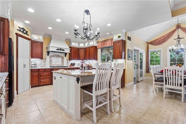 kitchen featuring light stone countertops, a notable chandelier, premium range hood, a kitchen bar, and a center island