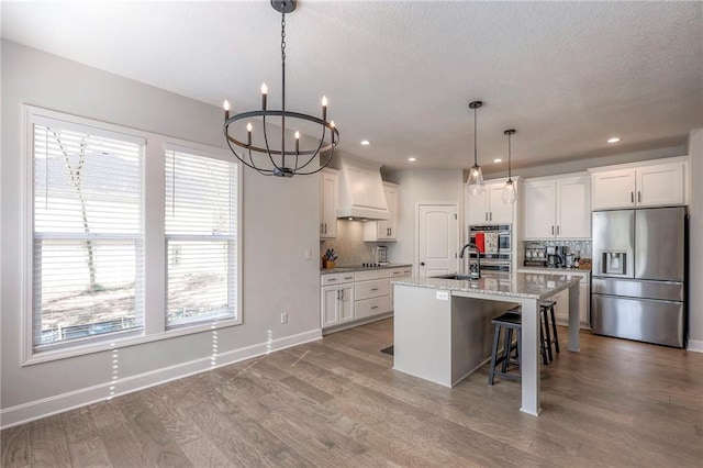 kitchen featuring white cabinets, custom exhaust hood, stainless steel appliances, and an island with sink