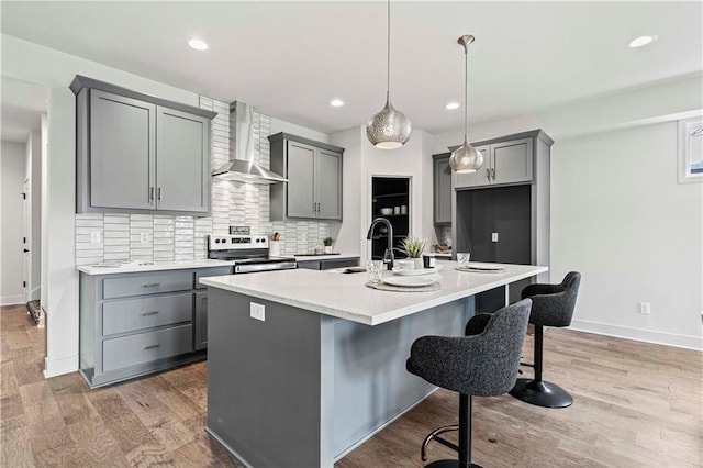 kitchen featuring gray cabinetry, wall chimney exhaust hood, decorative light fixtures, a kitchen island with sink, and electric range