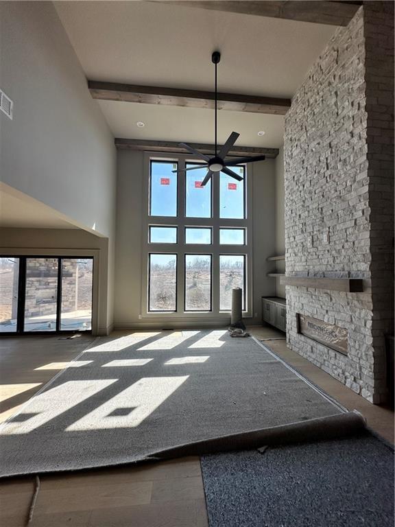 unfurnished living room featuring ceiling fan, beamed ceiling, a stone fireplace, a high ceiling, and wood finished floors