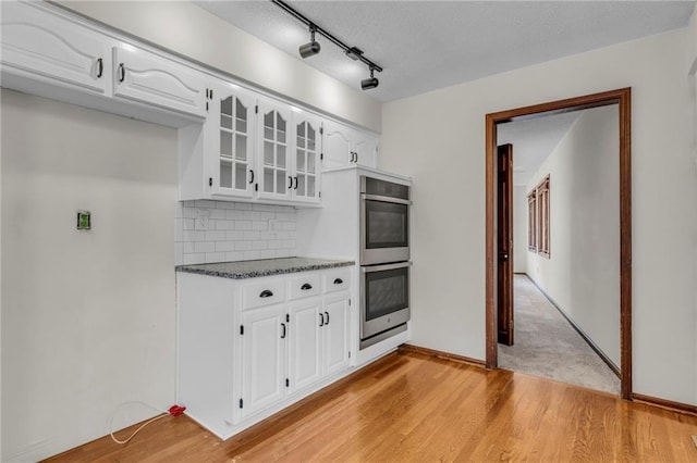 kitchen with white cabinets, stainless steel double oven, light wood-type flooring, dark stone counters, and tasteful backsplash