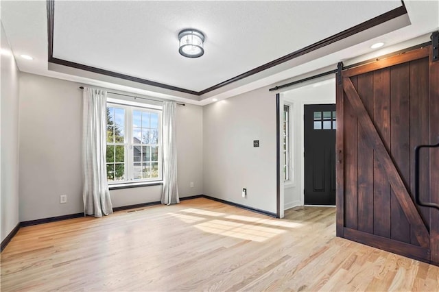 empty room with crown molding, a raised ceiling, light wood-style flooring, a barn door, and baseboards