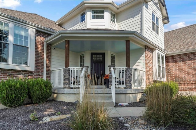 doorway to property with covered porch, brick siding, and roof with shingles