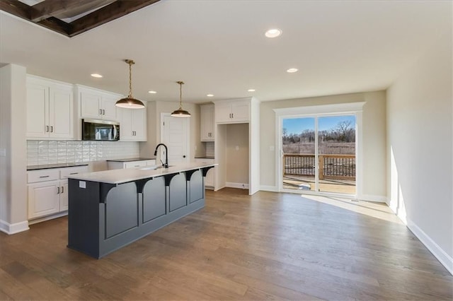 kitchen with tasteful backsplash, stainless steel microwave, a kitchen island with sink, white cabinetry, and a sink