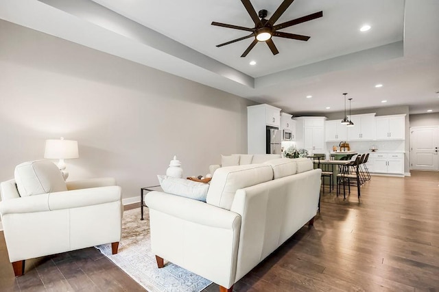 living area with a ceiling fan, a tray ceiling, baseboards, dark wood-style flooring, and recessed lighting