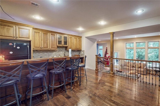 kitchen featuring decorative backsplash, dark wood-type flooring, black refrigerator, a kitchen bar, and decorative columns