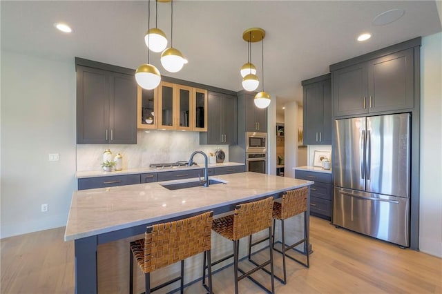 kitchen with light stone counters, stainless steel appliances, light hardwood / wood-style flooring, decorative light fixtures, and a kitchen island with sink