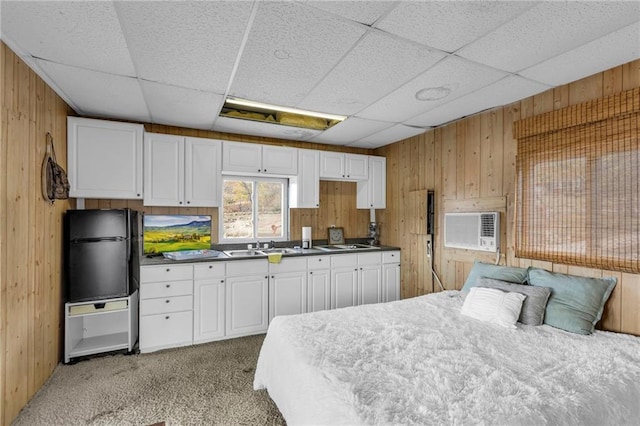 bedroom featuring wood walls, freestanding refrigerator, a drop ceiling, and light colored carpet