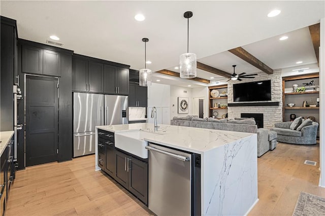 kitchen featuring appliances with stainless steel finishes, an island with sink, a brick fireplace, beam ceiling, and sink