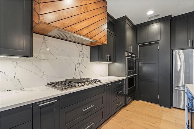 kitchen featuring light wood-type flooring, tasteful backsplash, custom range hood, stainless steel appliances, and light stone countertops