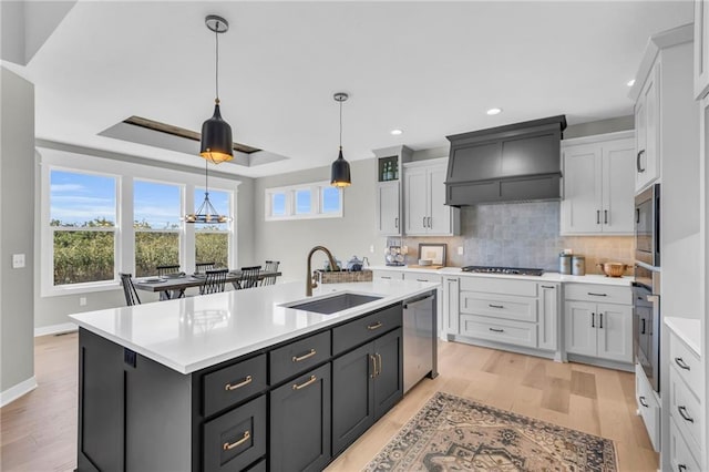 kitchen with a kitchen island with sink, custom range hood, stainless steel appliances, sink, and light hardwood / wood-style floors