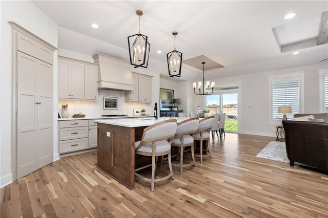 kitchen featuring light wood-type flooring, premium range hood, an island with sink, and decorative light fixtures