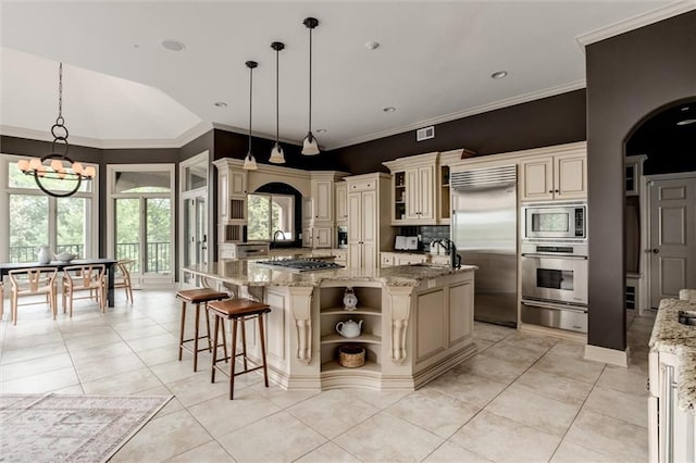 kitchen featuring light stone counters, an island with sink, built in appliances, decorative light fixtures, and cream cabinets