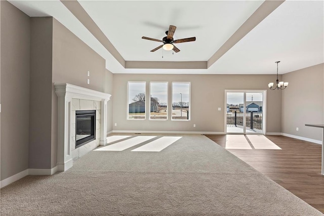 unfurnished living room with ceiling fan with notable chandelier, wood finished floors, baseboards, a tray ceiling, and a tiled fireplace