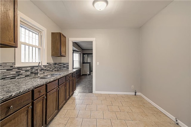 kitchen with decorative backsplash, a sink, dark brown cabinets, dark stone counters, and baseboards