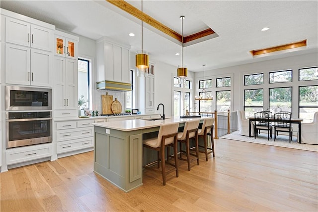 kitchen featuring light wood-type flooring, a kitchen island with sink, white cabinetry, appliances with stainless steel finishes, and decorative light fixtures