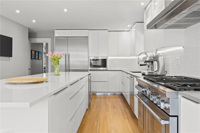 kitchen featuring tasteful backsplash, wall chimney exhaust hood, built in appliances, light hardwood / wood-style flooring, and white cabinetry