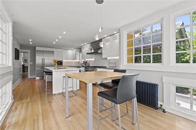 kitchen featuring white cabinetry, radiator heating unit, stainless steel built in refrigerator, decorative light fixtures, and a breakfast bar area