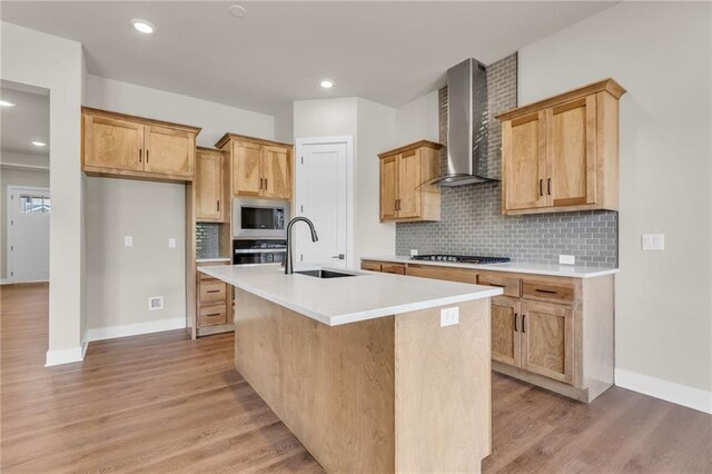 kitchen with appliances with stainless steel finishes, an island with sink, light wood-type flooring, sink, and wall chimney range hood