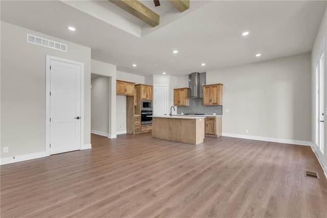 kitchen with an island with sink, tasteful backsplash, wall chimney exhaust hood, appliances with stainless steel finishes, and light wood-type flooring