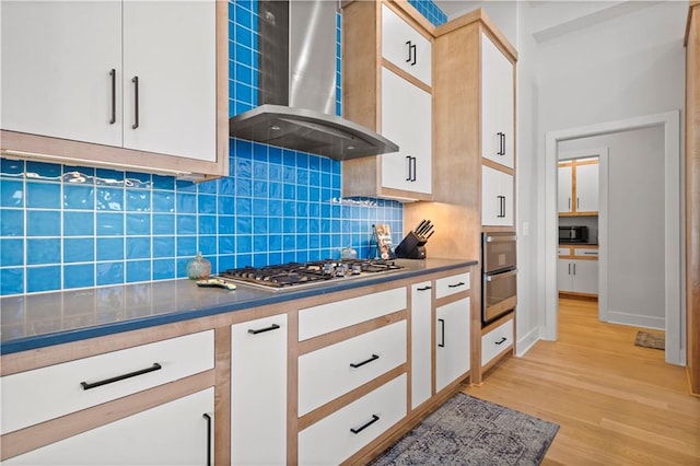 kitchen with backsplash, wall chimney exhaust hood, white cabinets, stainless steel gas stovetop, and light wood-type flooring