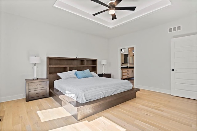 bedroom featuring a tray ceiling, light wood-type flooring, ceiling fan, and ensuite bathroom