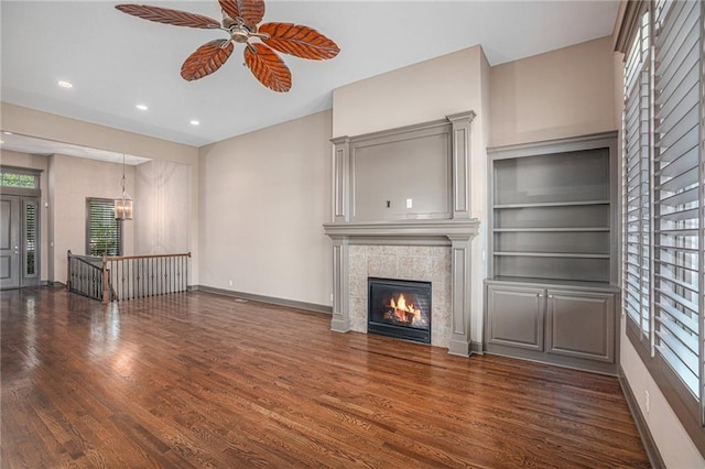 unfurnished living room featuring ceiling fan, a tiled fireplace, built in shelves, and dark hardwood / wood-style flooring