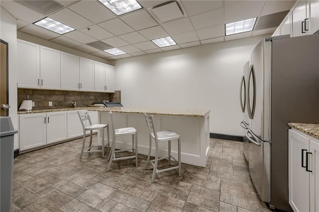 kitchen featuring decorative backsplash, a kitchen breakfast bar, light stone countertops, white cabinetry, and stainless steel refrigerator