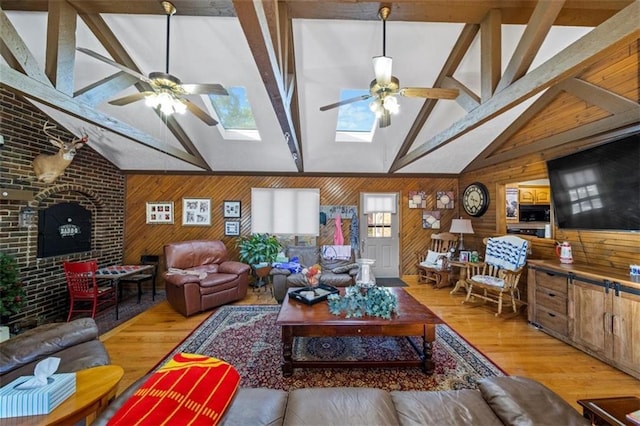 living room with wooden walls, a ceiling fan, vaulted ceiling with skylight, and light wood-style floors