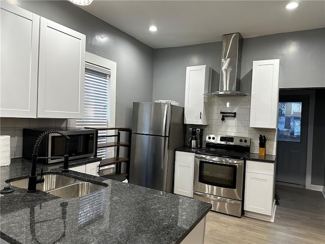 kitchen featuring wall chimney exhaust hood, white cabinetry, appliances with stainless steel finishes, dark stone countertops, and light hardwood / wood-style floors