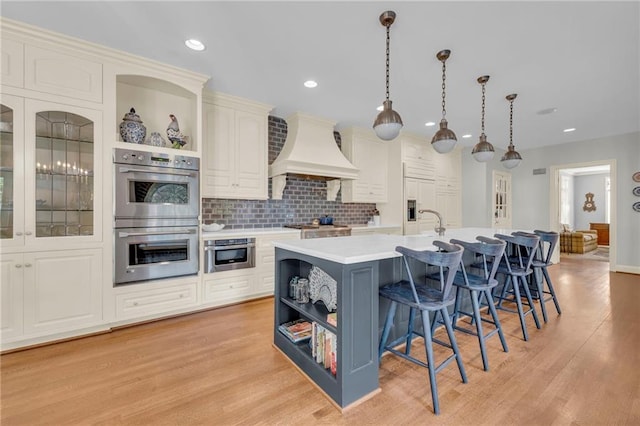 kitchen with a center island with sink, custom range hood, pendant lighting, light hardwood / wood-style floors, and stainless steel double oven