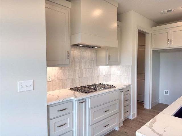kitchen featuring stainless steel gas cooktop, light stone counters, custom exhaust hood, light wood-type flooring, and backsplash
