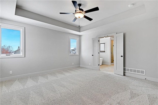 unfurnished bedroom featuring a tray ceiling, carpet, baseboards, ensuite bathroom, and a ceiling fan
