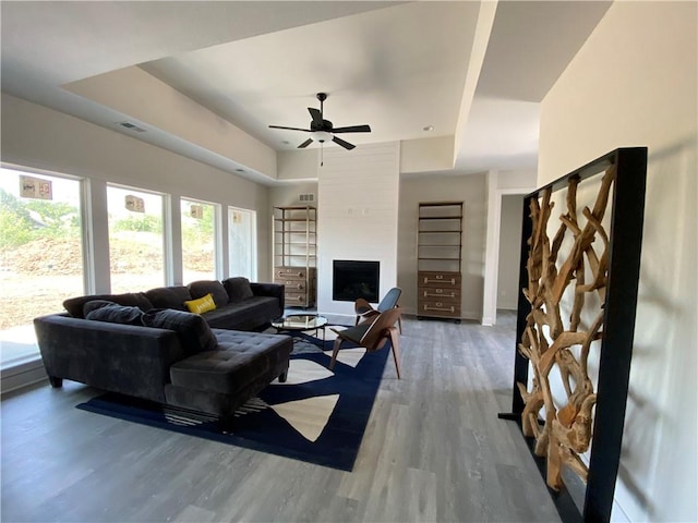 living room with wood-type flooring, a raised ceiling, ceiling fan, and a large fireplace