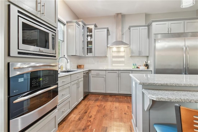 kitchen featuring wall chimney range hood, light stone counters, built in appliances, light hardwood / wood-style floors, and sink