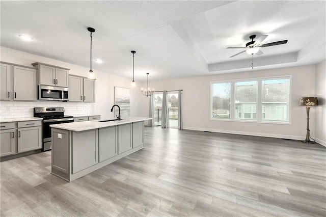 kitchen featuring hanging light fixtures, a raised ceiling, an island with sink, gray cabinets, and appliances with stainless steel finishes