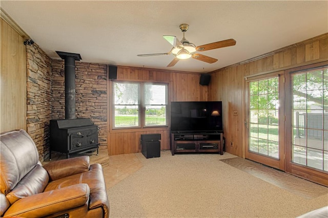 living room featuring wood walls, a wood stove, ceiling fan, crown molding, and carpet