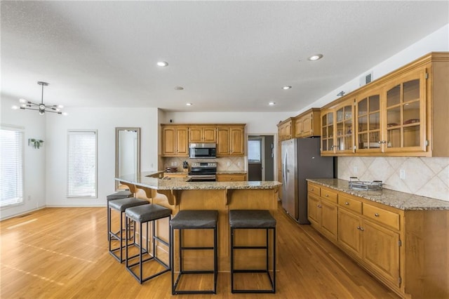 kitchen with stainless steel appliances, a center island with sink, a notable chandelier, tasteful backsplash, and light hardwood / wood-style flooring