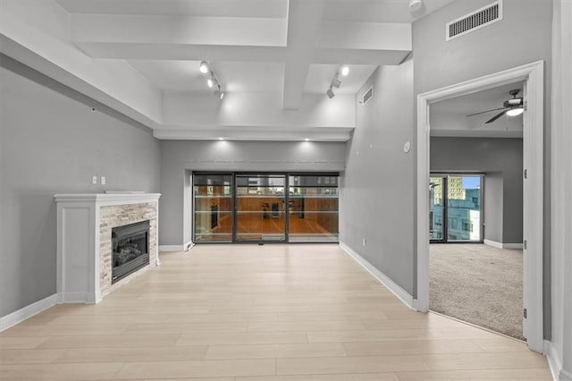 unfurnished living room featuring a stone fireplace, track lighting, beam ceiling, light colored carpet, and ceiling fan