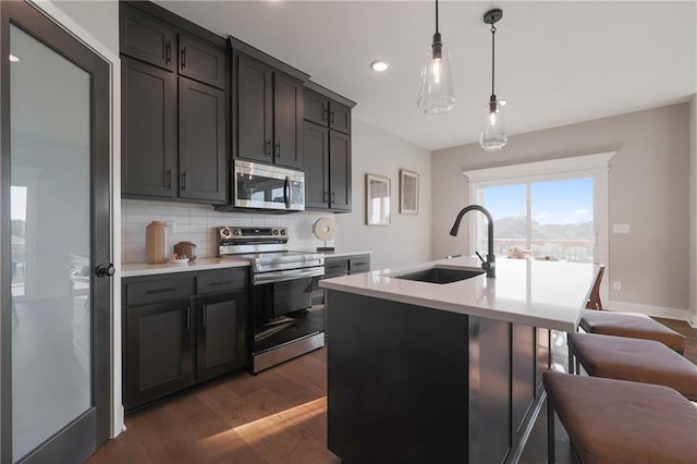 kitchen with dark wood-type flooring, a center island with sink, sink, decorative light fixtures, and stainless steel appliances