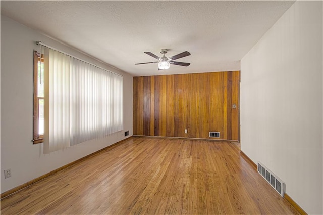 empty room with a textured ceiling, light hardwood / wood-style flooring, ceiling fan, and wooden walls