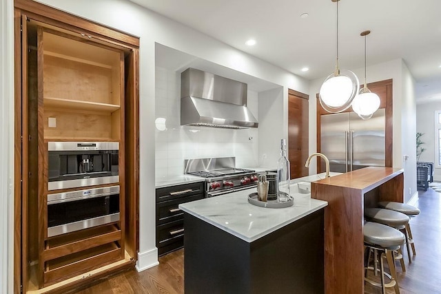 kitchen with wall chimney range hood, an island with sink, high quality appliances, decorative light fixtures, and dark wood-type flooring