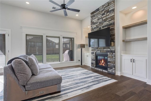 living room with dark wood-type flooring, ceiling fan, a stone fireplace, and built in shelves