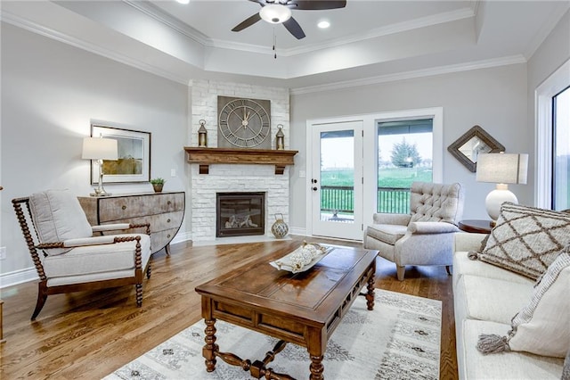 living room featuring hardwood / wood-style floors, ceiling fan, a raised ceiling, a stone fireplace, and crown molding