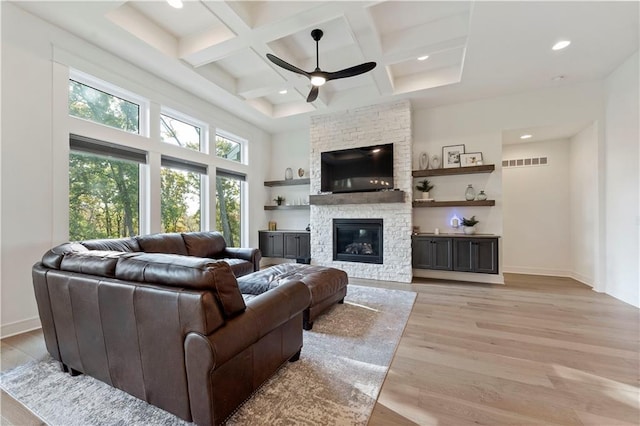 living room featuring light wood-type flooring, a stone fireplace, ceiling fan, coffered ceiling, and beamed ceiling