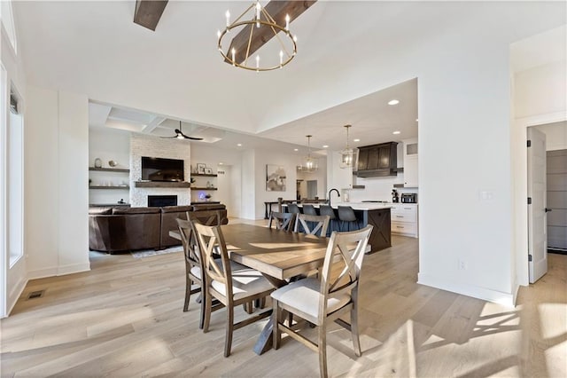 dining room with sink, light hardwood / wood-style flooring, a stone fireplace, and ceiling fan with notable chandelier