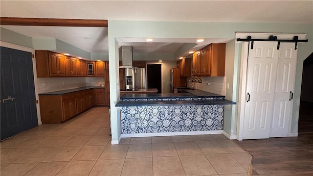 kitchen with a barn door, backsplash, wall chimney exhaust hood, stainless steel fridge with ice dispenser, and light tile patterned floors