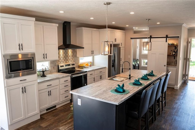 kitchen featuring a barn door, wall chimney exhaust hood, a center island with sink, and stainless steel appliances