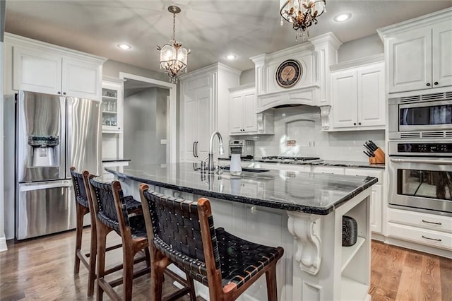 kitchen featuring light hardwood / wood-style flooring, a kitchen island with sink, pendant lighting, appliances with stainless steel finishes, and dark stone countertops